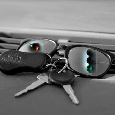 Sunglasses sit on dash of car. Reflecting in its eye pieces are traffic signals. Car keys lay in front of glasses.